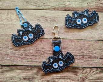 May include: Three black bat-shaped keychains with blue accents. The bats have white eyes and a sparkly black fabric. One keychain has a blue button and a silver ring.
