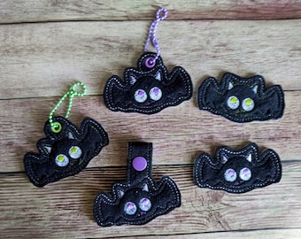 May include: Five black felt bat keychains with purple and white embroidered eyes. The bats have a 3D effect and are hanging on a wooden surface.