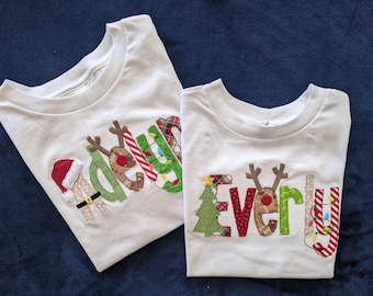 May include: Two white t-shirts with Christmas-themed appliques spelling out the names "Adelyn" and "Everly".