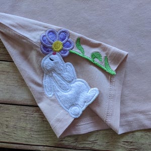 May include: A light pink t-shirt with a white bunny applique holding a purple flower with a yellow center and green stem. The bunny is embroidered with white thread. The t-shirt is folded, showing the bunny and flower design.