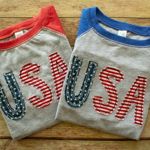 May include: Two grey t-shirts with red and blue raglan sleeves. The shirts have the word "USA" spelled out in large, colourful letters made of fabric.