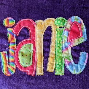 May include: A purple towel with the name "jane" spelled out in colorful fabric letters. Each letter is made from different patterned fabrics, including stripes, polka dots, and floral prints.