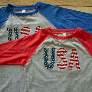 May include: Two grey, red and blue baseball t-shirts with the word "USA" in red, white and blue fabric on the front.