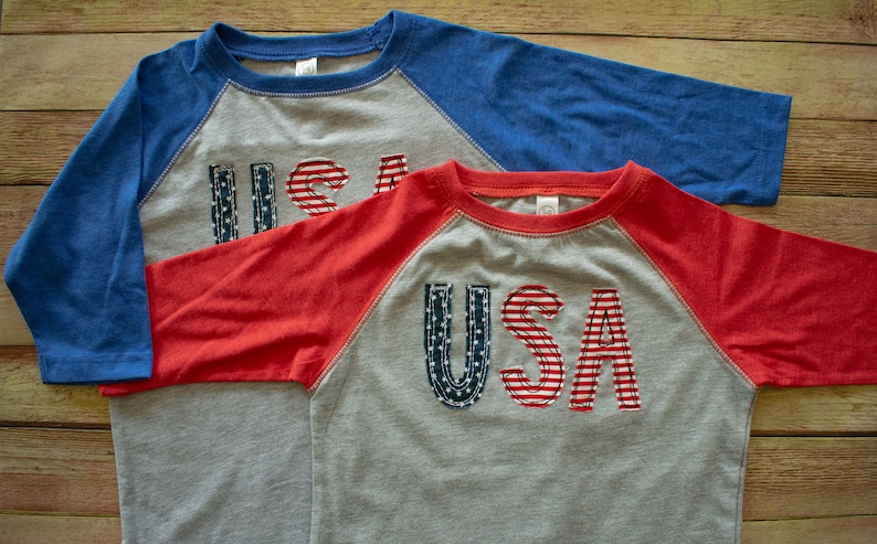 May include: Two grey, red and blue baseball t-shirts with the word "USA" in red, white and blue fabric on the front.
