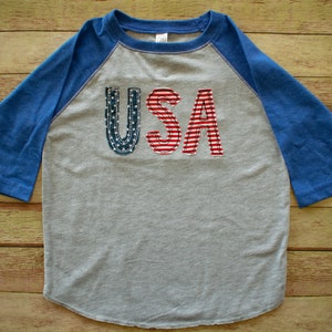 May include: A grey and blue baseball tee shirt with the word "USA" in red, white and blue fabric letters.
