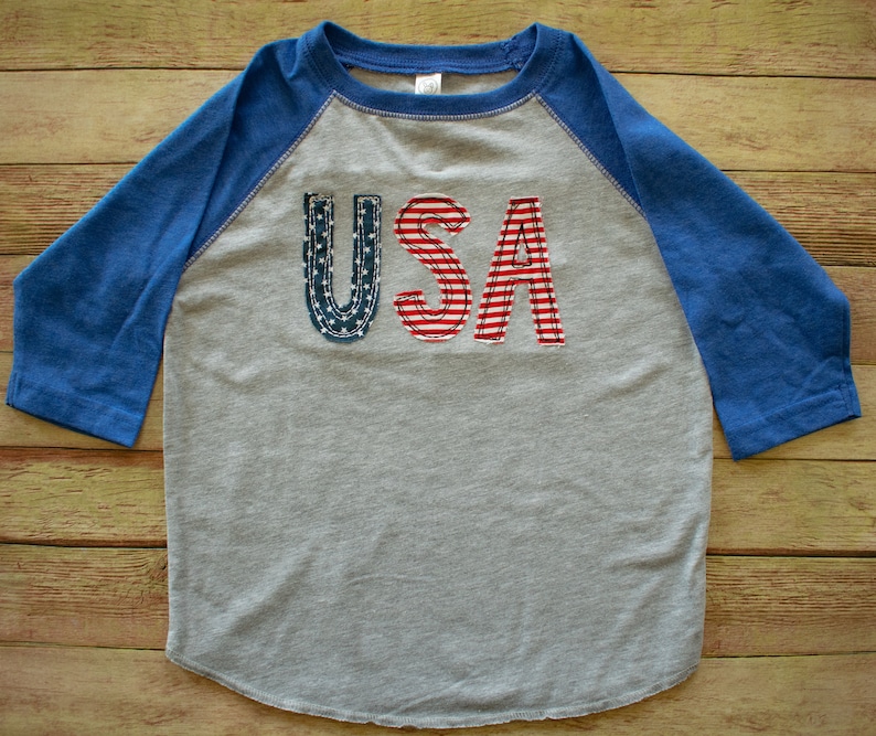 May include: A grey and blue baseball tee shirt with the word "USA" in red, white and blue fabric letters.