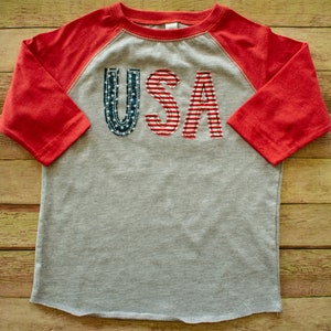 May include: A grey and red raglan t-shirt with the word "USA" in red, white and blue fabric letters.