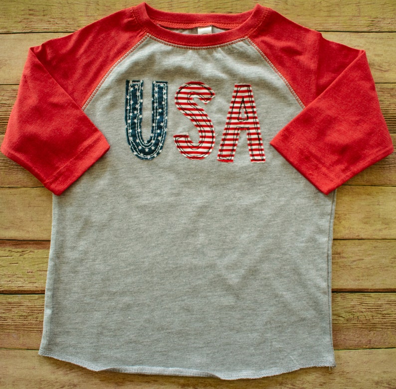 May include: A grey and red raglan t-shirt with the word "USA" in red, white and blue fabric letters.