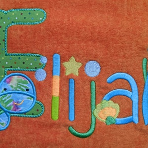 May include: A personalized towel with the name "Elijah" embroidered in blue and green. The towel features a green turtle with a blue shell and a blue jellyfish with green seaweed.