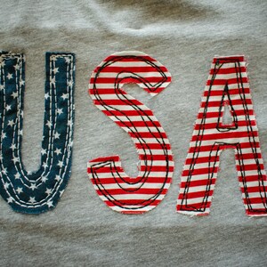 May include: A grey sweatshirt with the word "USA" sewn on in red, white and blue fabric. The letters are made of fabric with a striped pattern and a star pattern.