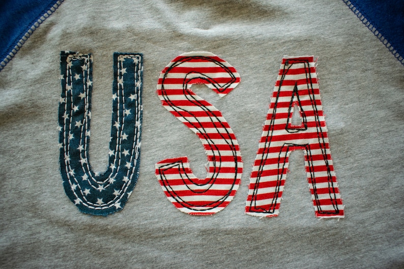 May include: A grey sweatshirt with the word "USA" sewn on in red, white and blue fabric. The letters are made of fabric with a striped pattern and a star pattern.