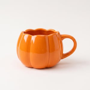 May include: A bright orange ceramic mug shaped like a pumpkin. The mug features a rounded, ribbed body with vertical indentations mimicking the appearance of a gourd, a wide circular opening at the top, and a curved handle on the right side.