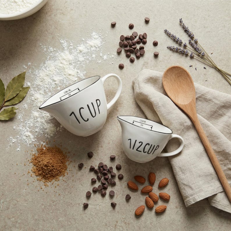 May include: Two white ceramic measuring jugs, marked "1 CUP" and "1/2 CUP", with black detailing. Ingredients such as chocolate chips, almonds, and flour are scattered around. A wooden spoon and lavender sprigs suggest baking.