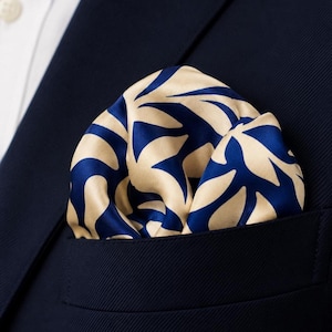 May include: A pocket square featuring a bold, repeating leaf pattern in deep navy blue and champagne gold. The fabric has a smooth, satin-like sheen and is folded neatly into the breast pocket of a dark navy blue suit jacket worn over a white shirt.