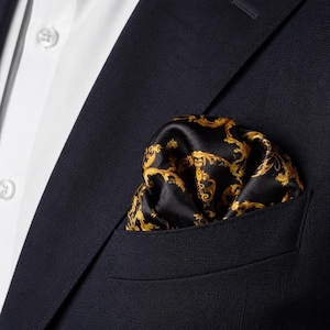 Op de afbeelding: Een zwart pochet met een goudkleurig, sierlijk krulpatroon in de borstzak van een donkerblauwe blazer, gedragen over een wit overhemd met zichtbare witte knopen op de kraag en de knoopsluiting aan de voorzijde van het kledingstuk.