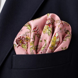 May include: A pink pocket square with a floral pattern featuring red, yellow, and green botanical illustrations, tucked into the breast pocket of a dark navy blue blazer. The fabric has a smooth, satin-like finish with visible folds.