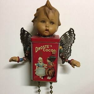 May include: A whimsical angel figurine made from repurposed materials. The angel has a porcelain doll head, a red tin can body with the words "Droste's Coco" printed on it, and wooden clogs for feet. The angel has wings made from metal and is adorned with beads and other embellishments.