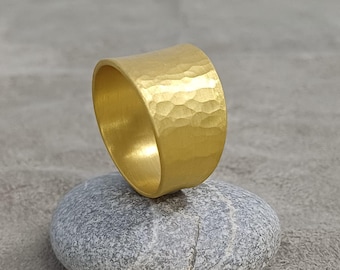 May include: A wide, gold-colored ring with a hammered texture. The ring is displayed on a gray stone against a gray background. The ring's surface reflects light, highlighting its textured design. The ring appears to be made of metal.