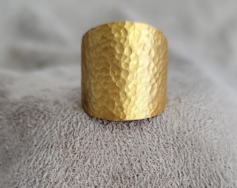 May include: A wide gold-toned ring featuring a distinct hammered texture across its entire outer surface. The metal has a matte finish with small, concave indentations that create a repetitive, organic pattern throughout the band's exterior.