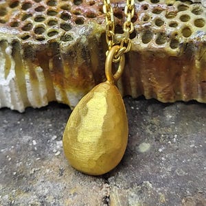 May include: A gold-toned pendant shaped like a teardrop with a hammered, textured surface. The pendant is suspended from a matching gold-toned metal chain link necklace. The teardrop charm features a small circular bail at the top for attachment.