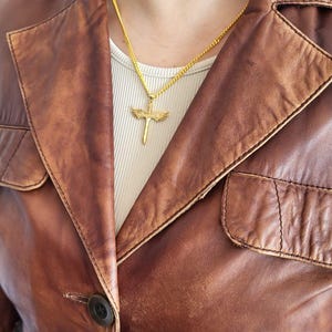 May include: A gold-toned necklace featuring a pendant shaped like a sword with outstretched wings. The pendant hangs from a thin gold-toned chain, resting against a white ribbed shirt worn underneath a brown leather jacket with a collared neckline.