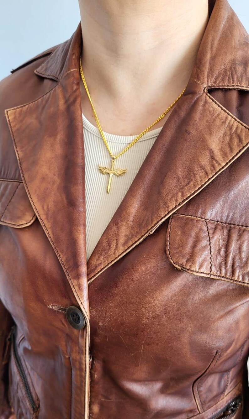 May include: A gold-toned necklace featuring a pendant shaped like a sword with outstretched wings. The pendant hangs from a thin gold-toned chain, resting against a white ribbed shirt worn underneath a brown leather jacket with a collared neckline.