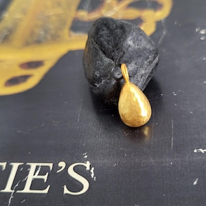 May include: A small, gold-coloured pendant shaped like a teardrop with a textured, hammered surface. The pendant features a small circular loop at the top for attachment. It is resting against a dark, rough, irregularly shaped stone object.
