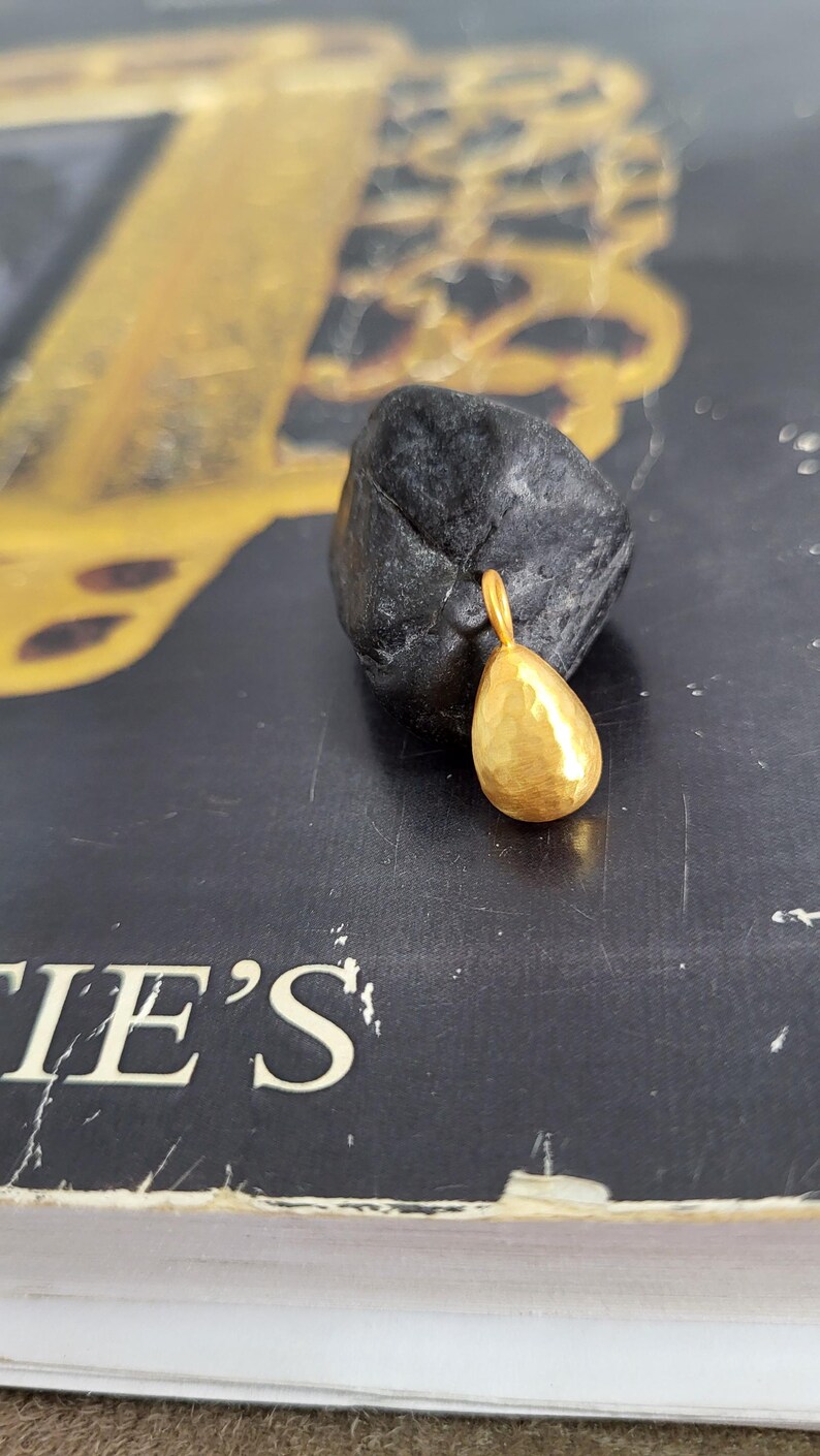 May include: A small, gold-coloured pendant shaped like a teardrop with a textured, hammered surface. The pendant features a small circular loop at the top for attachment. It is resting against a dark, rough, irregularly shaped stone object.