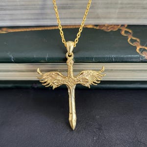 May include: A gold-toned necklace featuring a pendant shaped like a sword with outstretched wings. The sword handle is detailed with intricate engravings, and the blade points downward. The pendant hangs from a matching gold-toned chain link.