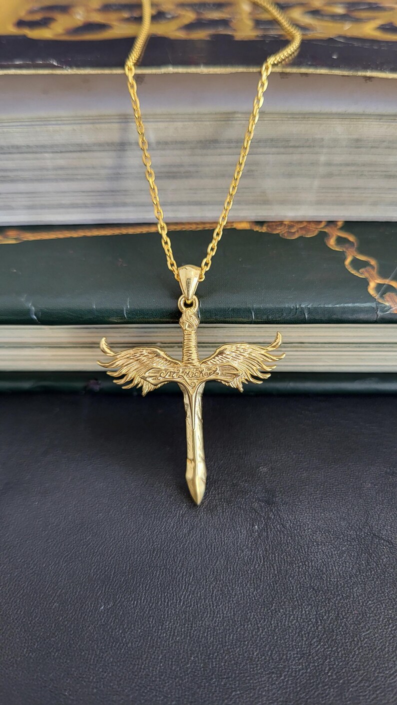May include: A gold-toned necklace featuring a pendant shaped like a sword with outstretched wings. The sword handle is detailed with intricate engravings, and the blade points downward. The pendant hangs from a matching gold-toned chain link.