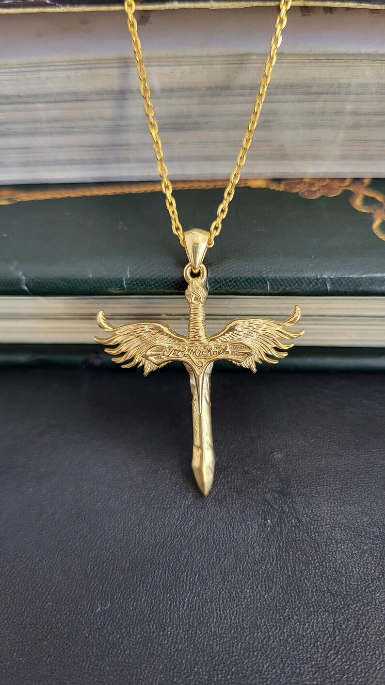 May include: A gold-toned necklace featuring a pendant shaped like a sword with detailed wings extending from the hilt. The center of the cross-guard is engraved with the text St. Michael. The pendant hangs from a matching gold-toned chain link.