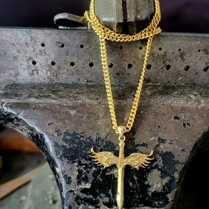May include: A gold-toned necklace featuring a pendant shaped like a sword with outstretched wings. The pendant is suspended from a classic curb chain. The sword design includes detailed feather textures on the wings and a pointed blade tip.