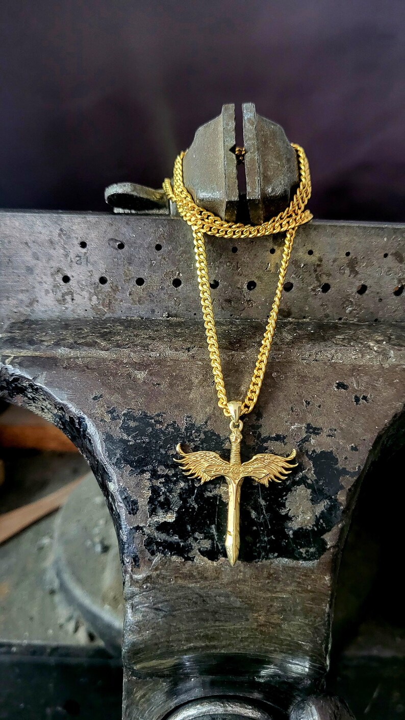 May include: A gold-toned necklace featuring a pendant shaped like a sword with outstretched wings. The pendant is suspended from a classic curb chain. The sword design includes detailed feather textures on the wings and a pointed blade tip.