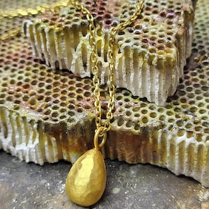 May include: A gold-coloured necklace featuring a teardrop-shaped pendant with a hammered, textured surface. The pendant is suspended from a delicate gold-coloured metal chain with small, uniform oval links, creating a simple and refined jewellery piece.