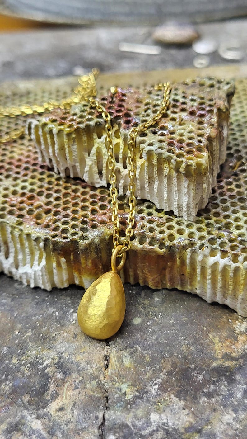 May include: A gold-coloured necklace featuring a teardrop-shaped pendant with a hammered, textured surface. The pendant is suspended from a delicate gold-coloured metal chain with small, uniform oval links, creating a simple and refined jewellery piece.
