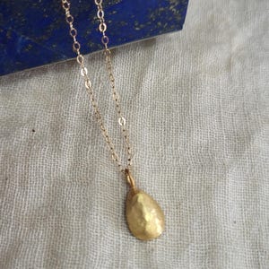 May include: A delicate gold necklace featuring a small, hammered, teardrop-shaped pendant suspended from a fine, polished gold chain. The pendant has a textured, matte finish that catches the light, creating a subtle, organic appearance.