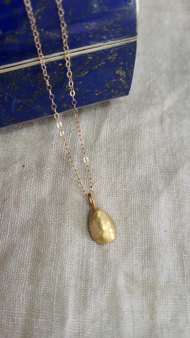 May include: A delicate gold necklace featuring a small, hammered, teardrop-shaped pendant suspended from a fine, polished gold chain. The pendant has a textured, matte finish that catches the light, creating a subtle, organic appearance.
