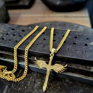 May include: A gold-toned metal necklace featuring a pendant shaped like a sword with outstretched wings. The pendant is attached to a classic curb chain. The design includes detailed feather textures on the wings and a pointed sword blade.