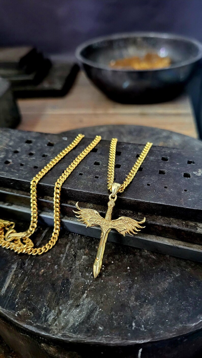May include: A gold-toned metal necklace featuring a pendant shaped like a sword with outstretched wings. The pendant is attached to a classic curb chain. The design includes detailed feather textures on the wings and a pointed sword blade.