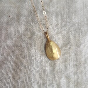 May include: A gold-toned necklace featuring a delicate chain and a single teardrop-shaped pendant. The pendant has a textured, hammered surface that reflects light, suspended from a simple metal link chain that connects to the top of the charm.