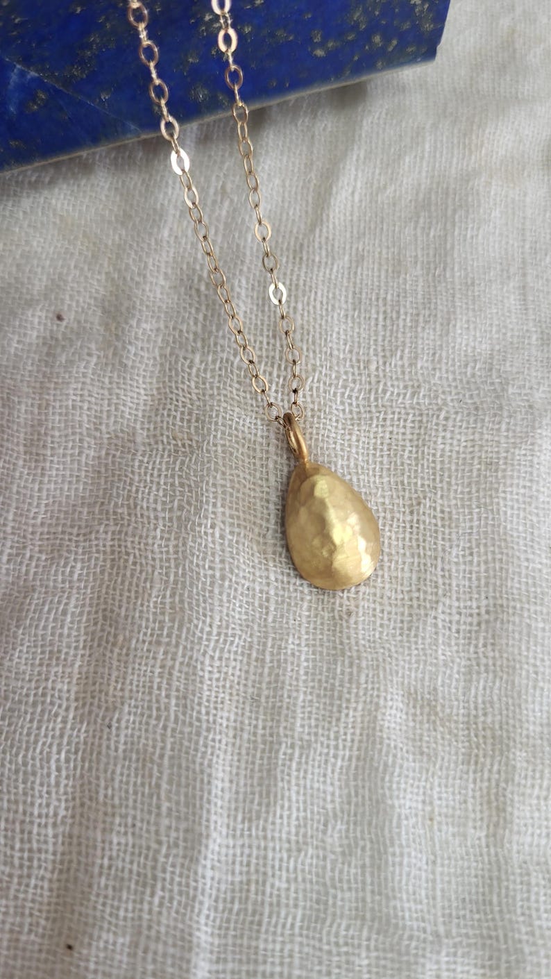 May include: A gold-toned necklace featuring a delicate chain and a single teardrop-shaped pendant. The pendant has a textured, hammered surface that reflects light, suspended from a simple metal link chain that connects to the top of the charm.