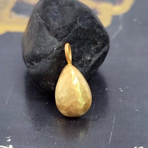 May include: A gold-toned pendant in a teardrop shape with a hammered, textured surface. The pendant features a small, circular bail at the top for attachment. The metal has a matte, brushed finish that catches the light across its uneven surface.