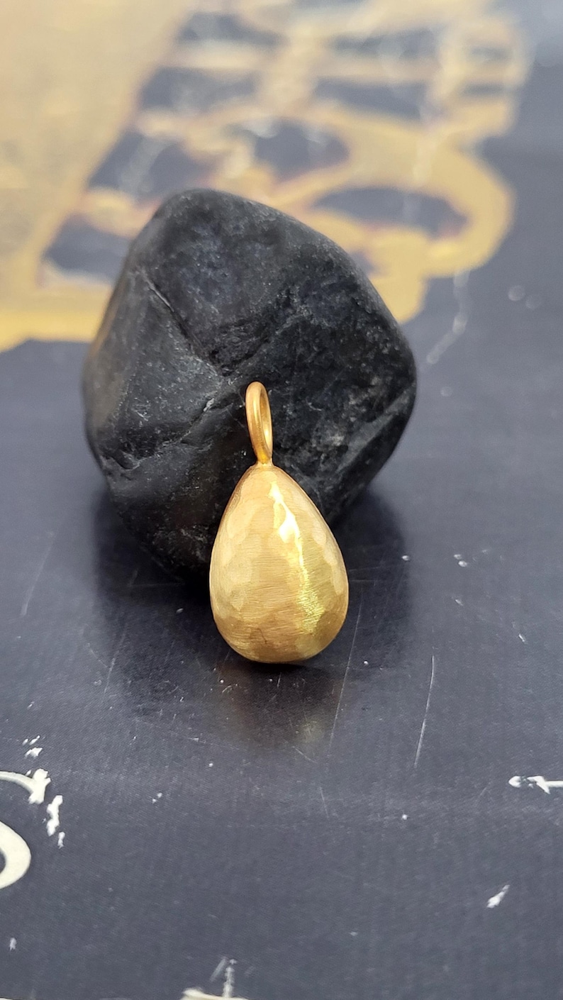 May include: A gold-toned pendant in a teardrop shape with a hammered, textured surface. The pendant features a small, circular bail at the top for attachment. The metal has a matte, brushed finish that catches the light across its uneven surface.