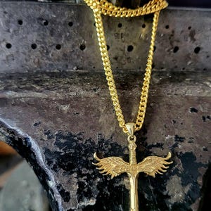May include: A gold-toned necklace featuring a pendant shaped like a sword with detailed wings extending from the hilt. The pendant hangs from a gold-toned curb chain, showcasing a metallic finish and intricate sculpted textures on the wings and blade.