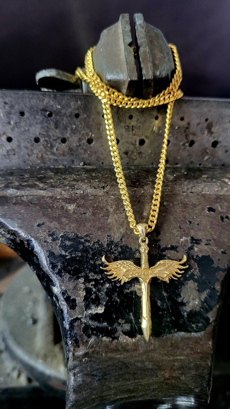 May include: A gold-toned necklace featuring a pendant shaped like a sword with detailed wings extending from the hilt. The pendant hangs from a gold-toned curb chain, showcasing a metallic finish and intricate sculpted textures on the wings and blade.