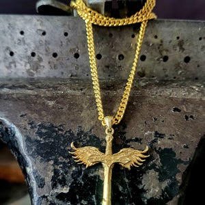 May include: A gold-toned necklace featuring a pendant shaped like a sword with outstretched wings. The pendant is suspended from a matching gold-toned curb chain. The sword design includes detailed feather textures on the wings and a pointed blade.