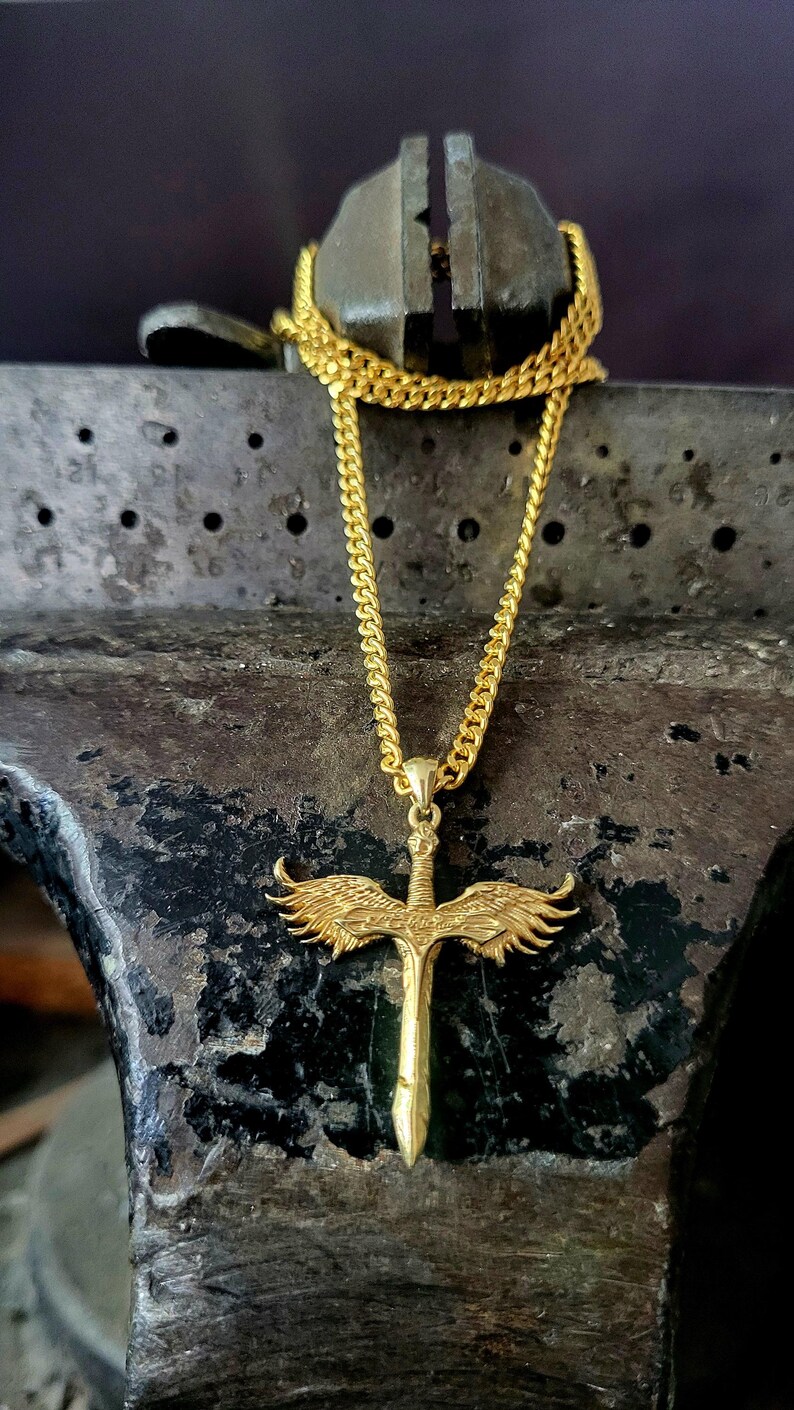 May include: A gold-toned necklace featuring a pendant shaped like a sword with outstretched wings. The pendant is suspended from a matching gold-toned curb chain. The sword design includes detailed feather textures on the wings and a pointed blade.