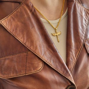 May include: A gold-toned metal necklace featuring a cross pendant with detailed wings extending from the sides. The pendant hangs from a thin gold-toned chain, resting against a beige ribbed top worn underneath a brown leather jacket with a collar.