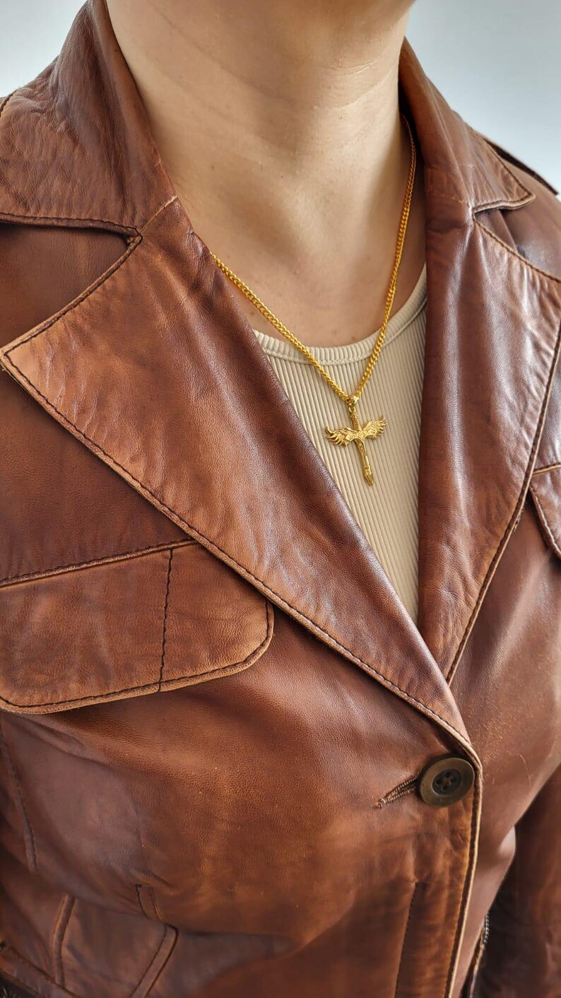 May include: A gold-toned metal necklace featuring a cross pendant with detailed wings extending from the sides. The pendant hangs from a thin gold-toned chain, resting against a beige ribbed top worn underneath a brown leather jacket with a collar.