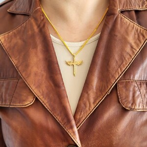 May include: A gold-toned necklace featuring a pendant shaped like a sword with outstretched wings. The pendant hangs from a delicate gold-toned chain, resting against a light-colored ribbed top worn underneath a brown leather jacket with a button.